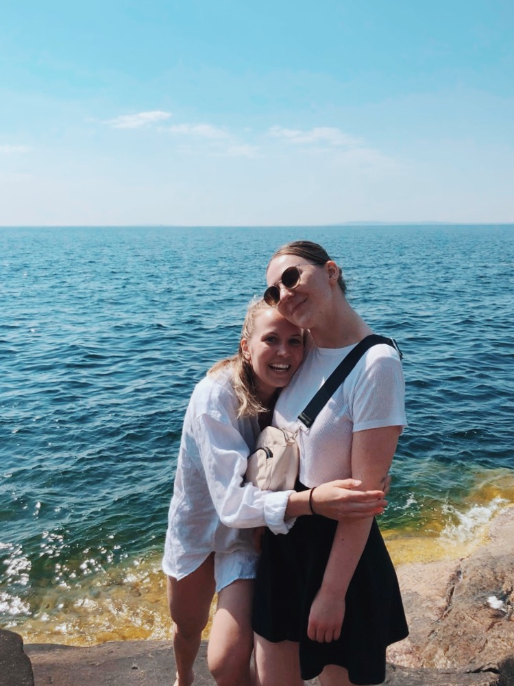 Två kvinnor står på en klippa med vatten bakom sig och håller om varandra.